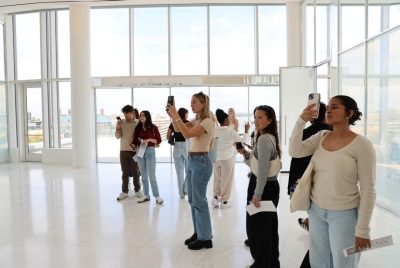 A group of students, some of whom are taking photos with cell phones, stands inside the interior of a building in front of a wall of windows