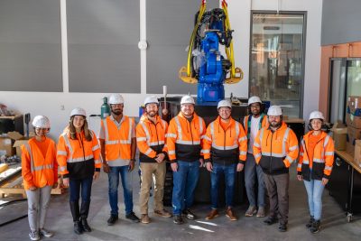 A group of people in reflective orange shirts and hardhats stand in front of large 3D printer with robotic arm.