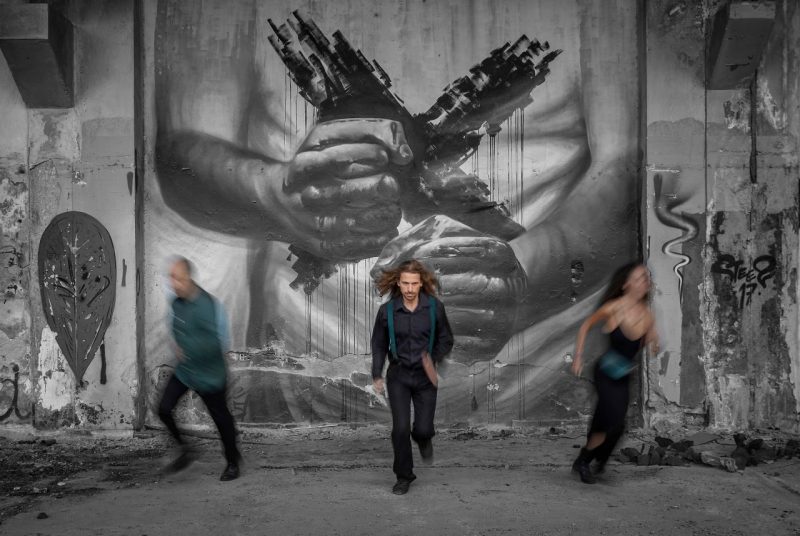 Three people run toward the camera in front of a large black-and-white mural of two clenched hands holding broken structures that resemble city buildings. The person in the center is sharply focused, wearing dark clothes with teal suspenders, while the figures on either side are blurred in motion. 