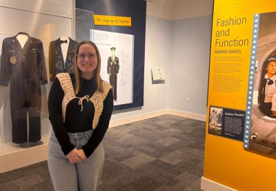 Juliette Good stands in front of glass cases holding uniforms and walls displaying information about the history of Navy uniforms.