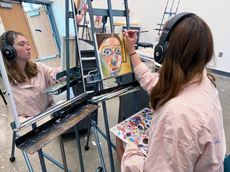 Student looking in mirror painting self portrait with headphones on